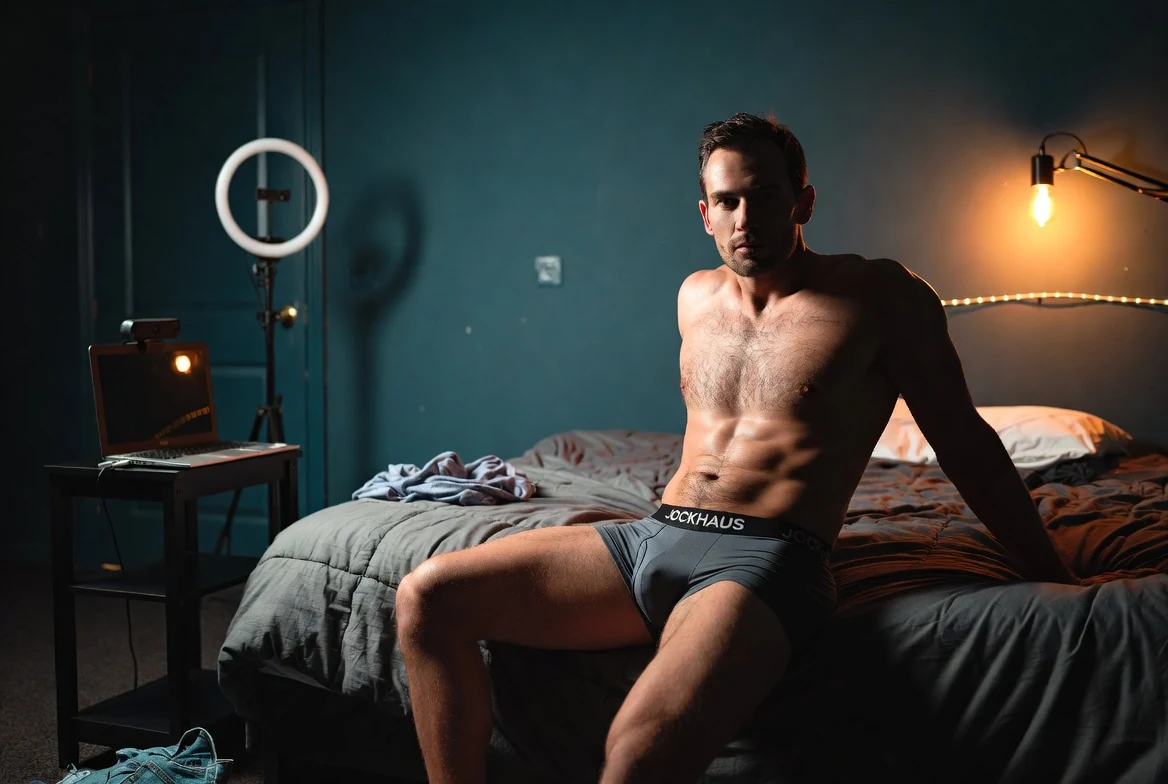 Adult male performer seated on a bed beside a laptop and ring light